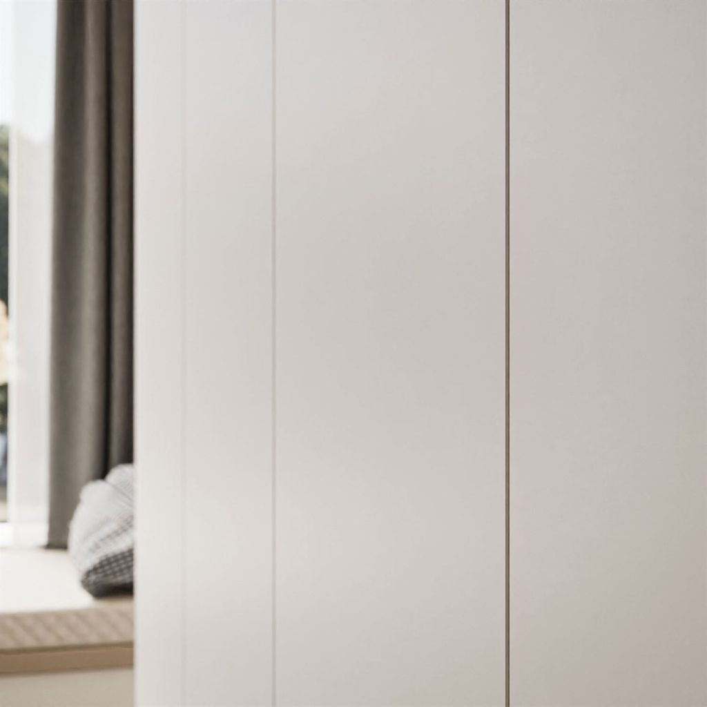 A minimalist interior scene with a large white wardrobe dominating the frame. To the left, a glimpse of a window with grey curtains and a cushioned window seat with a patterned throw.