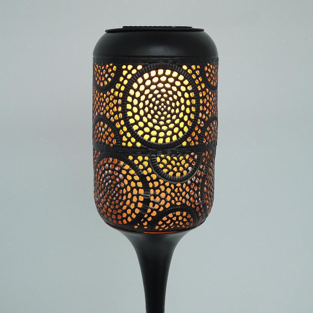 A close-up view of a black solar-powered garden light with intricate cut-out patterns that emit a warm, orange glow.