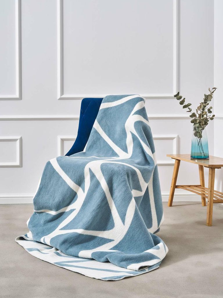 A blue and white geometric patterned blanket draped over a chair in a room with white paneled walls and a small wooden side table with a vase of greenery.