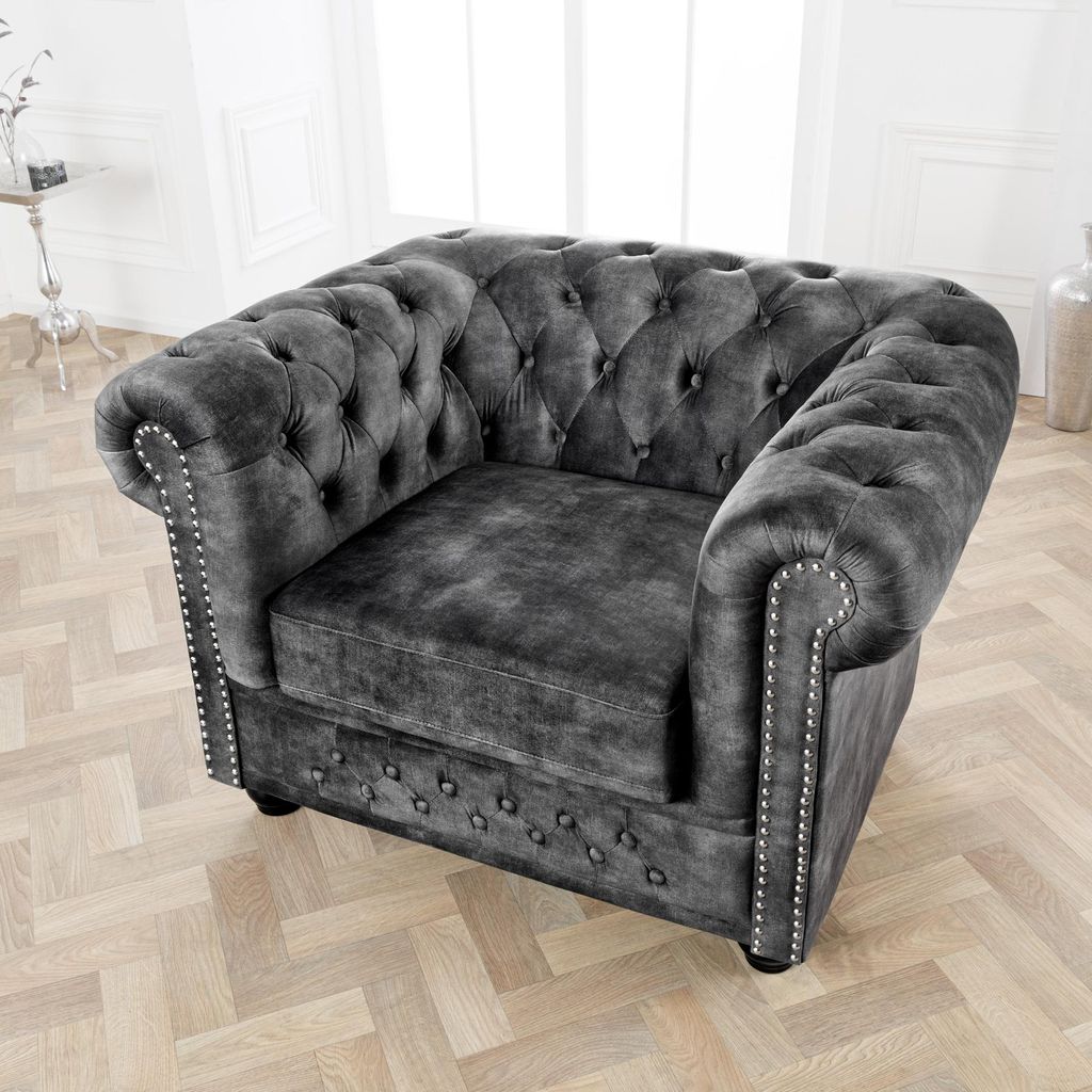 A dark gray velvet armchair with button tufting and silver nailhead trim, placed on a light wood floor in a well-lit room.
