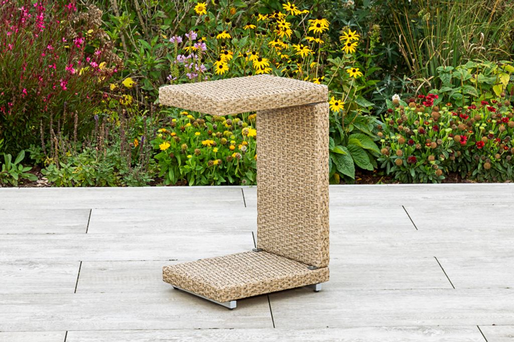 A C-shaped side table made of woven material sits on a patio with a garden in the background.