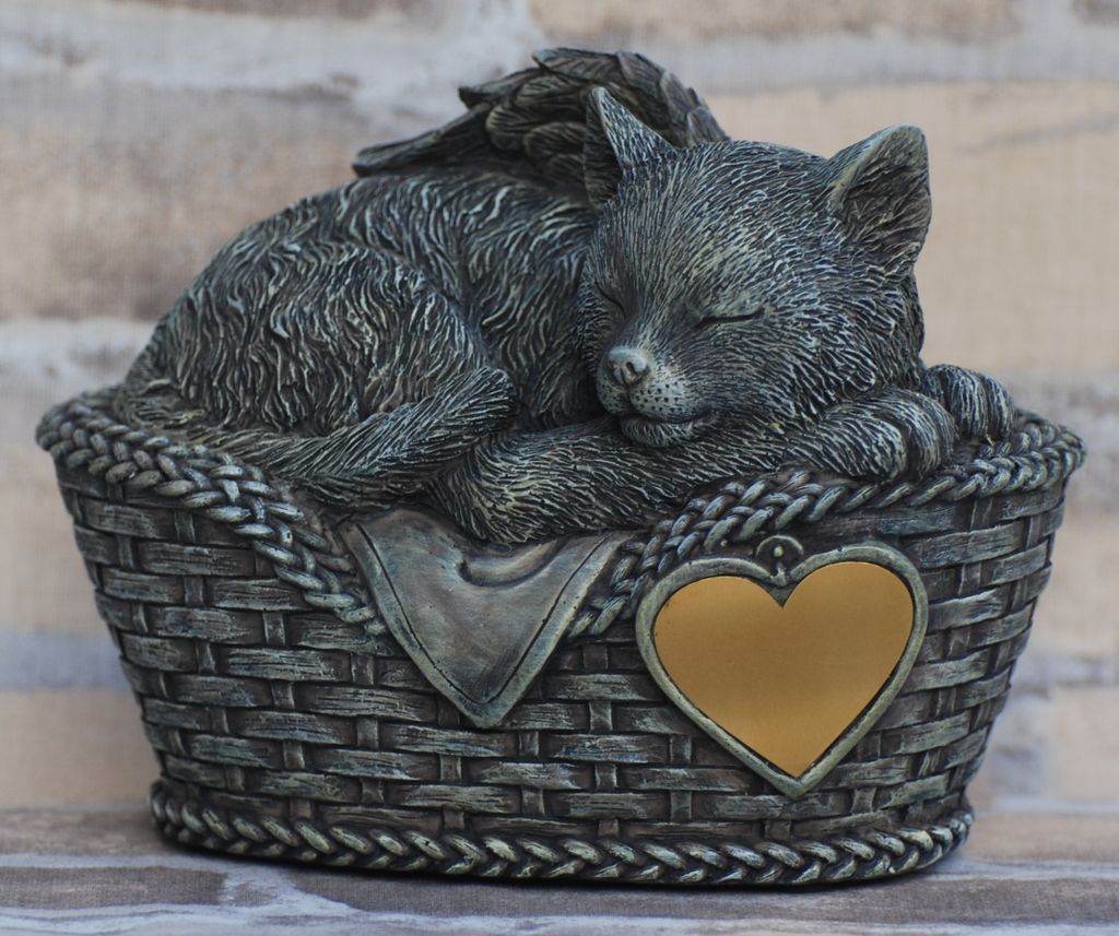 A sleeping cat figurine with wings, resting in a basket, with a heart-shaped plaque.