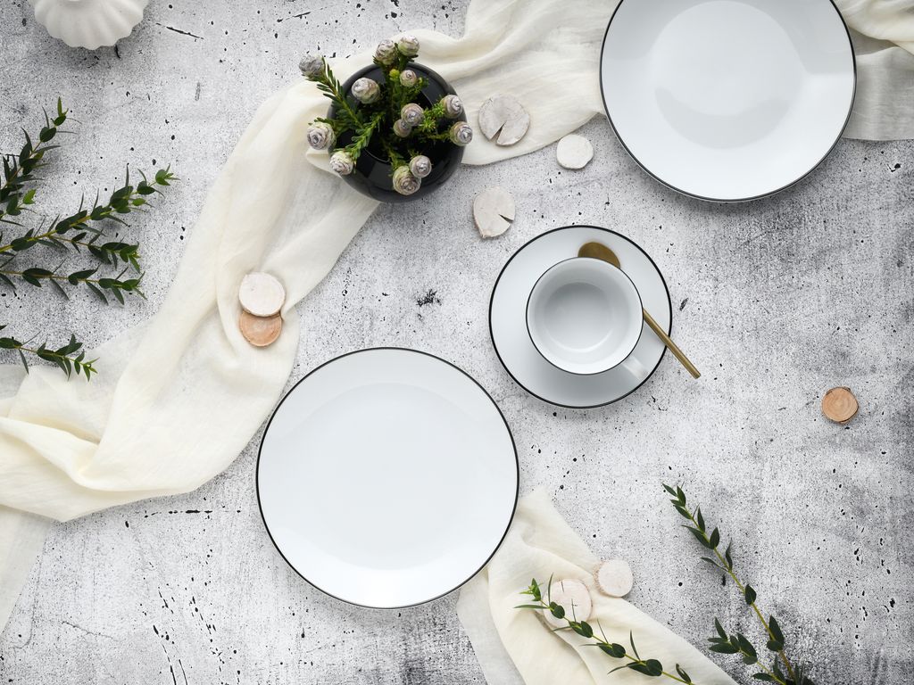 A flat lay of a white porcelain coffee service set with black trim, arranged on a textured gray surface with a cream-colored fabric runner and some greenery.