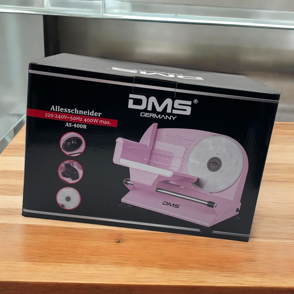 A pink meat slicer is shown on its packaging box, which is placed on a wooden surface.