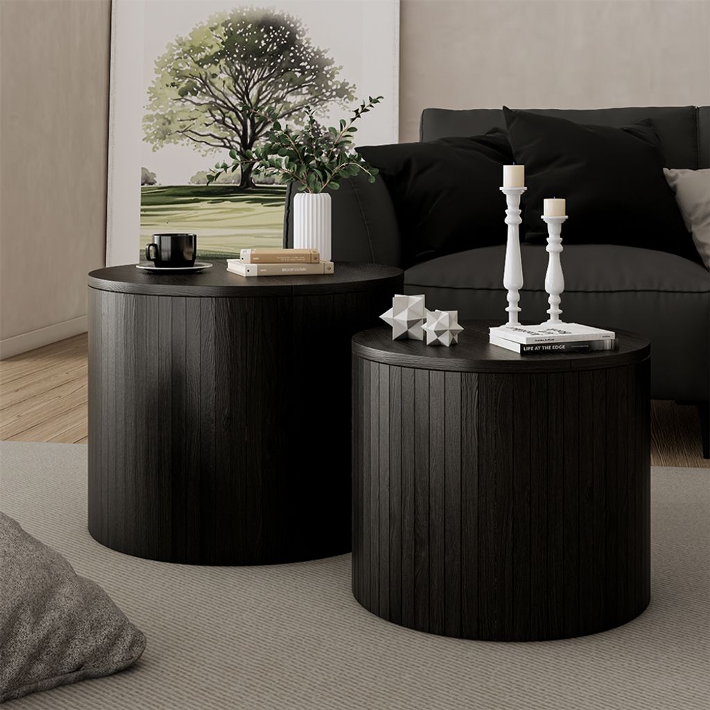 Two round black coffee tables with ribbed sides are placed on a gray rug in a living room setting. A painting of a tree hangs on the wall behind the tables, and a sofa with black cushions is visible in the background.