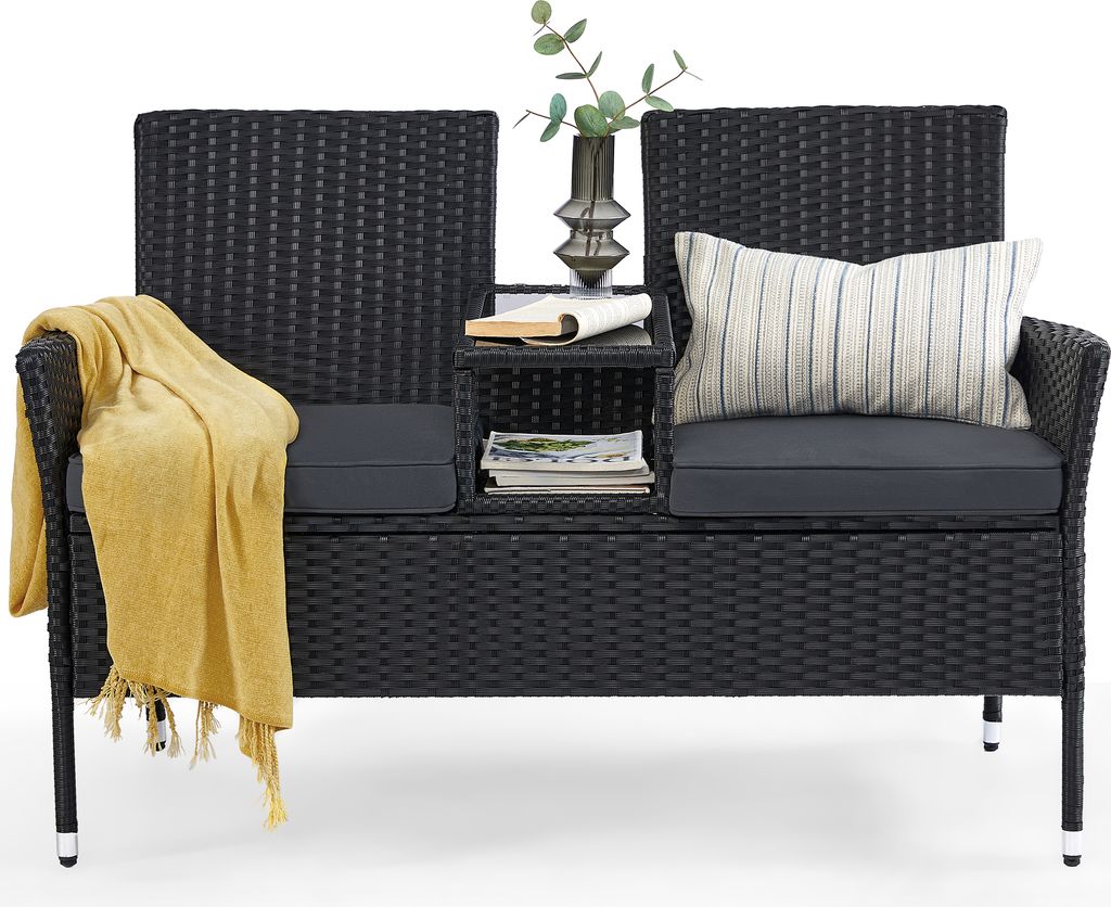 A black rattan bench with gray cushions, a yellow throw blanket, a pillow, books, and a vase with greenery.
