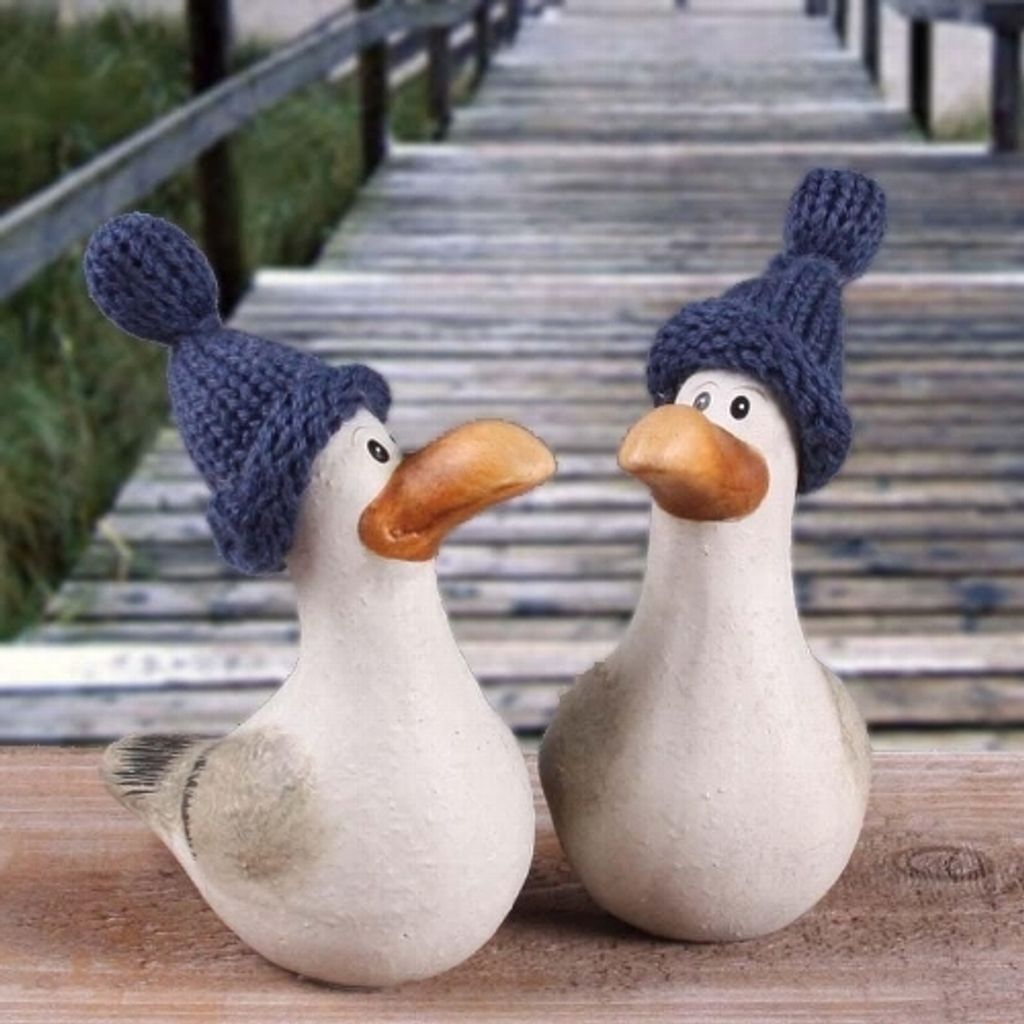 Two decorative seagulls wearing knitted blue hats stand on a wooden surface in front of a wooden boardwalk.