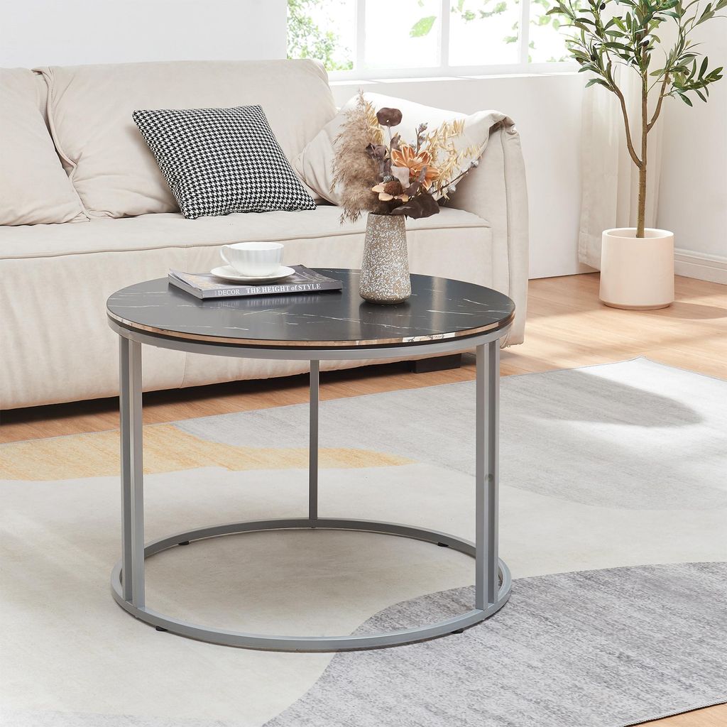 A round coffee table with a marble top and metal legs is placed on a rug in front of a sofa. A vase with dried flowers and a book with a cup of tea are on the table.