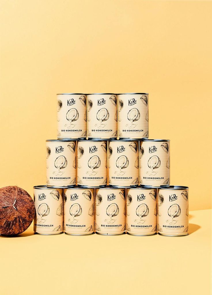 A pyramid stack of 12 cans of coconut milk, with a coconut half in the foreground.