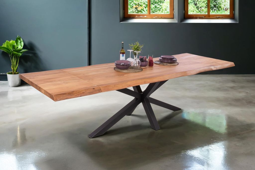 A wooden dining table with a live edge and a black metal spider leg base is set in a modern dining room with a dark gray wall and a concrete floor. Two windows with greenery visible are in the background.