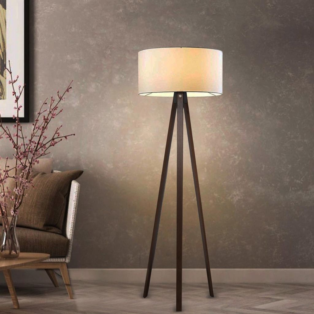 A floor lamp with a tripod base and a cream-colored shade is shown in a living room setting next to a sofa and a small table.