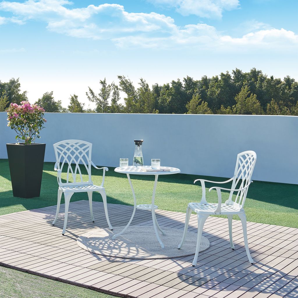 A white bistro set with a table and two chairs on a wooden deck with a potted plant and a wall in the background.