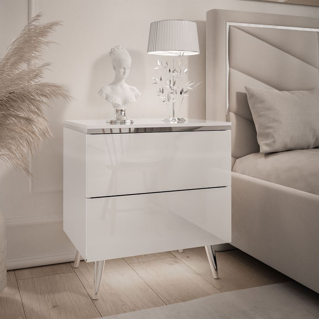 A white, high-gloss nightstand with chrome legs, decorated with a bust and a lamp, next to a bed.