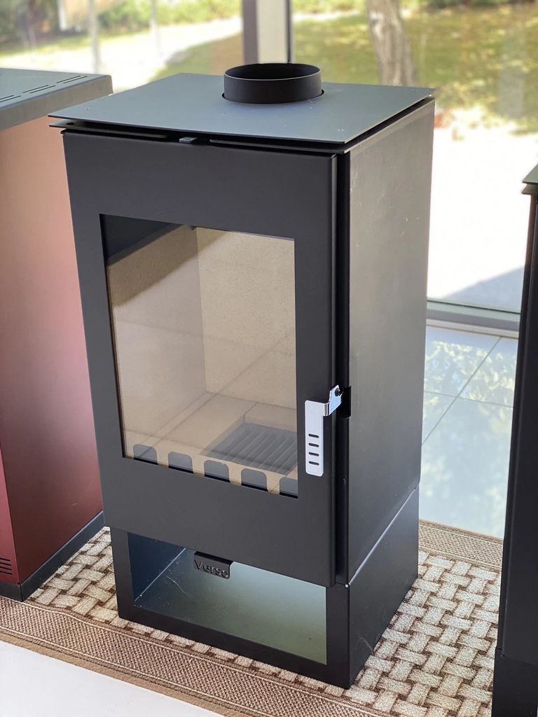 A black wood-burning stove with a glass door is displayed indoors, possibly in a showroom or home.
