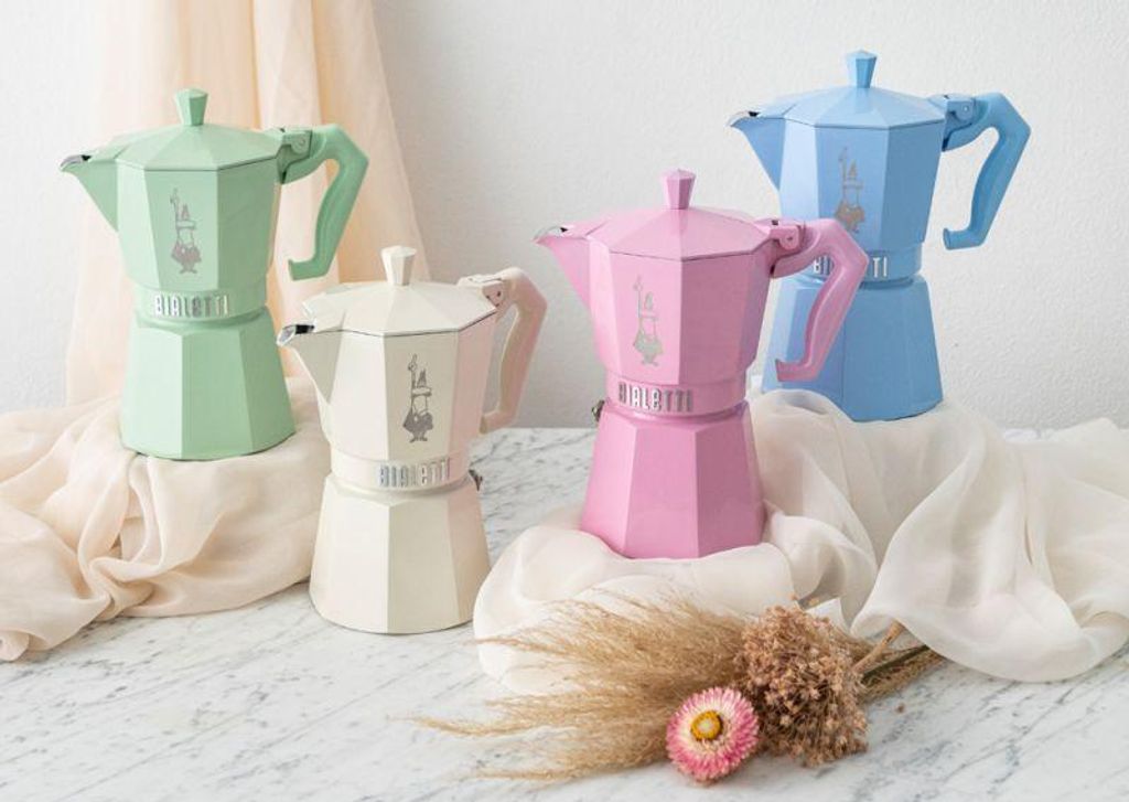 Four Bialetti Moka Express coffee makers in pastel colors (green, cream, pink, blue) arranged on a surface with draped fabric and dried flowers.