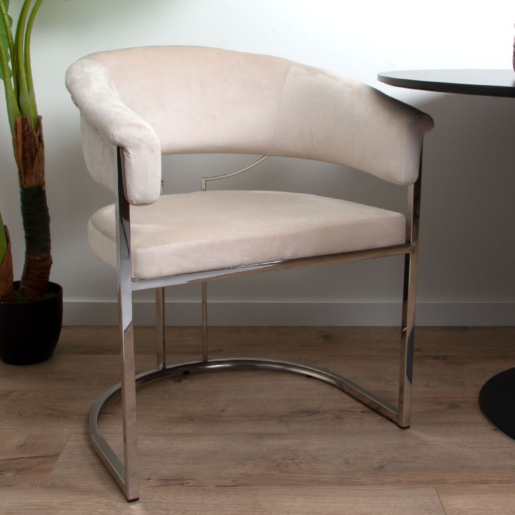 A beige velvet dining chair with a silver metal frame is shown in a room with a plant and a table.