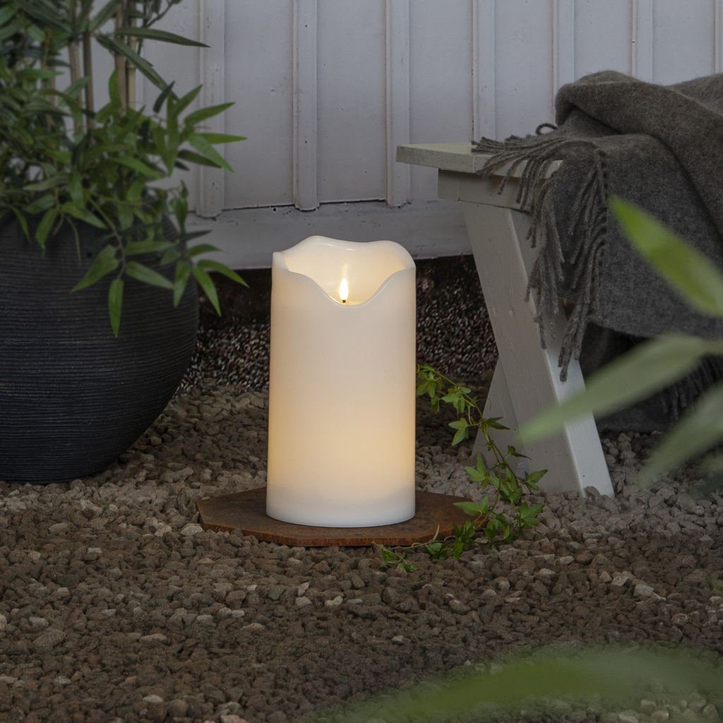 An LED candle is placed on gravel next to a potted plant and a bench with a blanket.