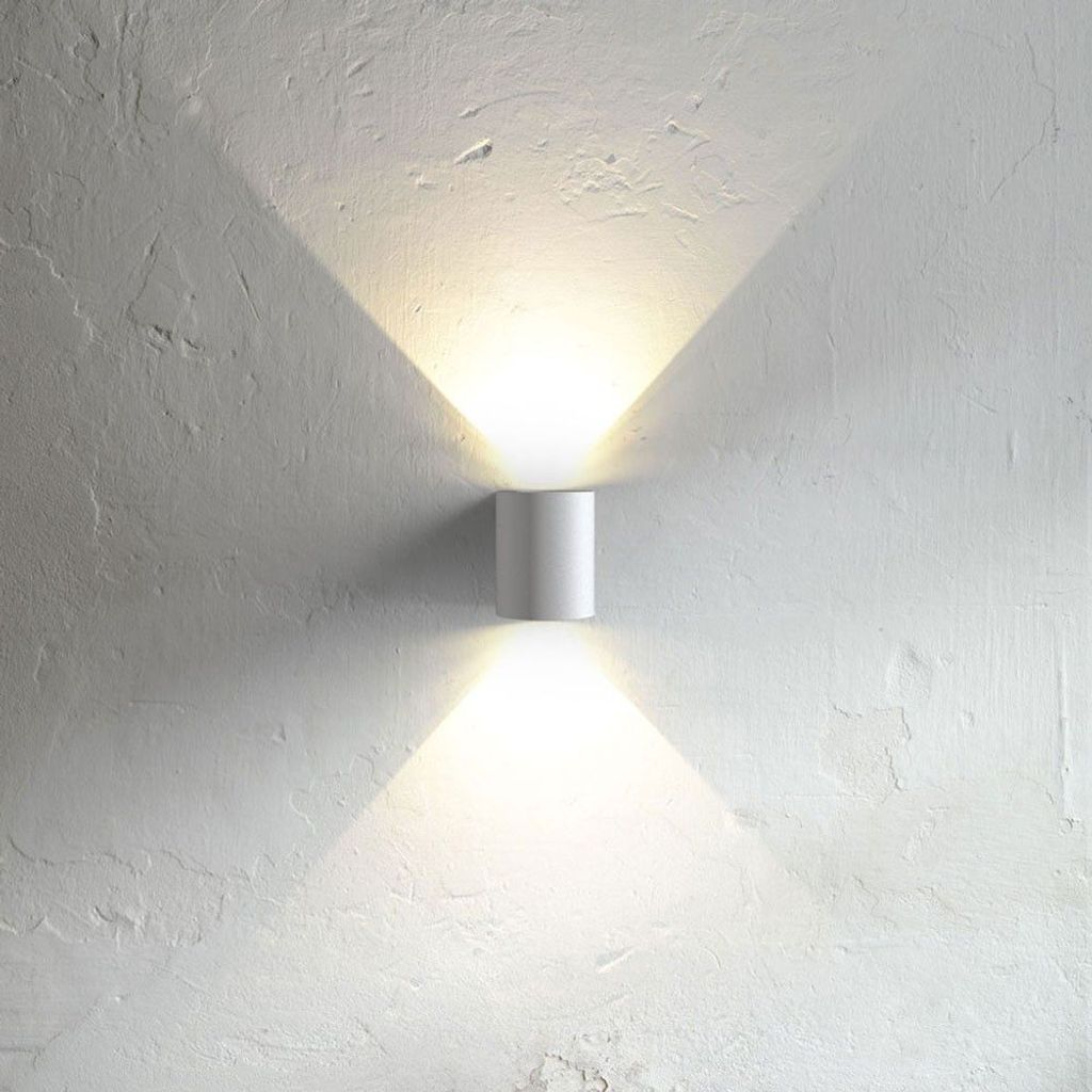 A white cylindrical wall lamp mounted on a textured white wall, casting light upwards and downwards.