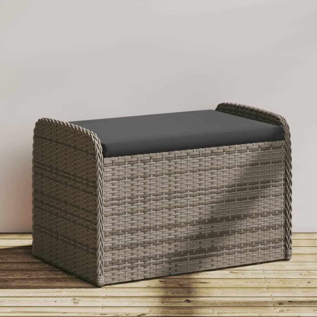 A grey poly rattan storage bench with a dark grey cushion is placed on a wooden deck against a light grey wall.