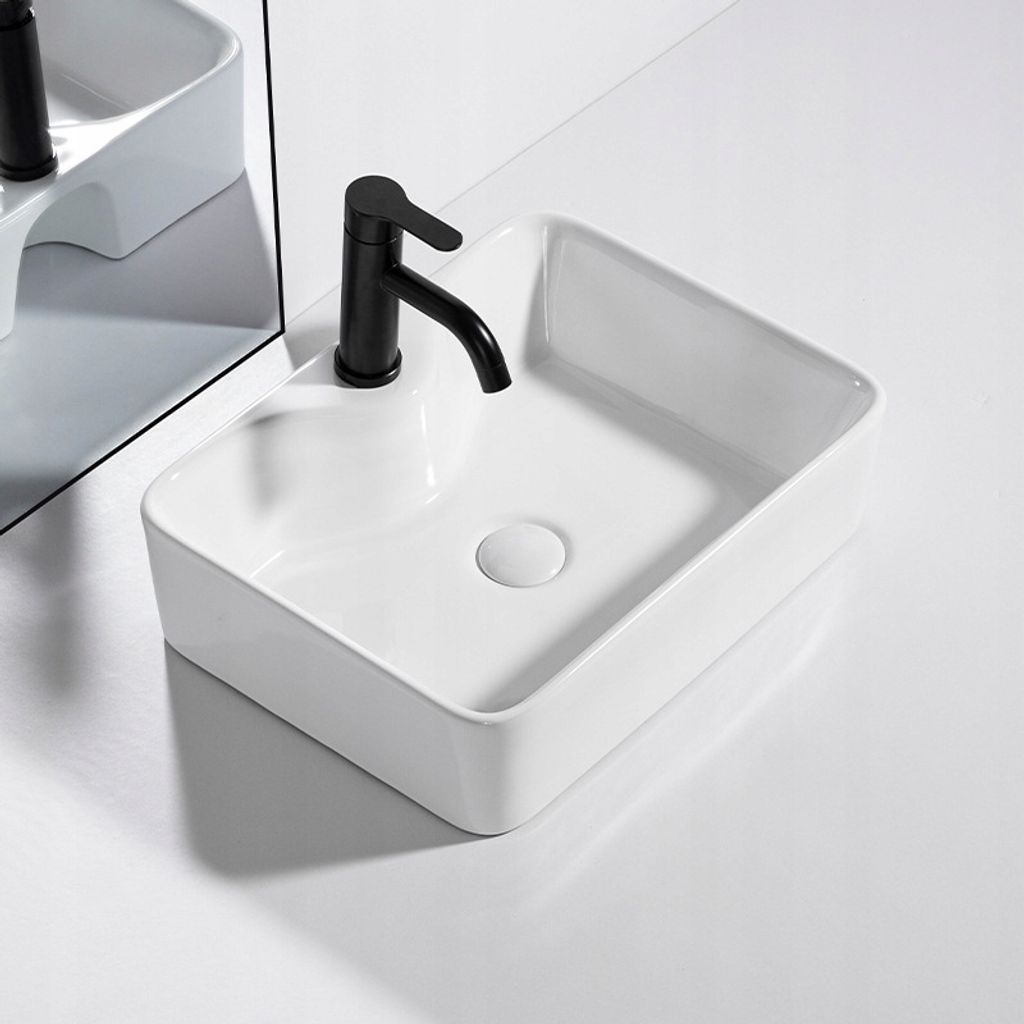 A white ceramic sink with a black faucet is shown on a white surface, with a mirror reflecting the sink and faucet.