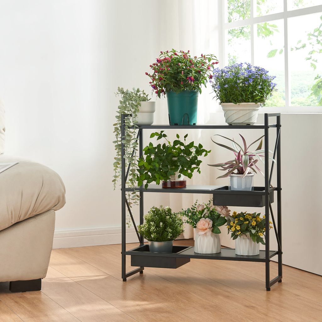 A black metal plant stand with multiple shelves filled with various potted plants, placed indoors near a window and a sofa.