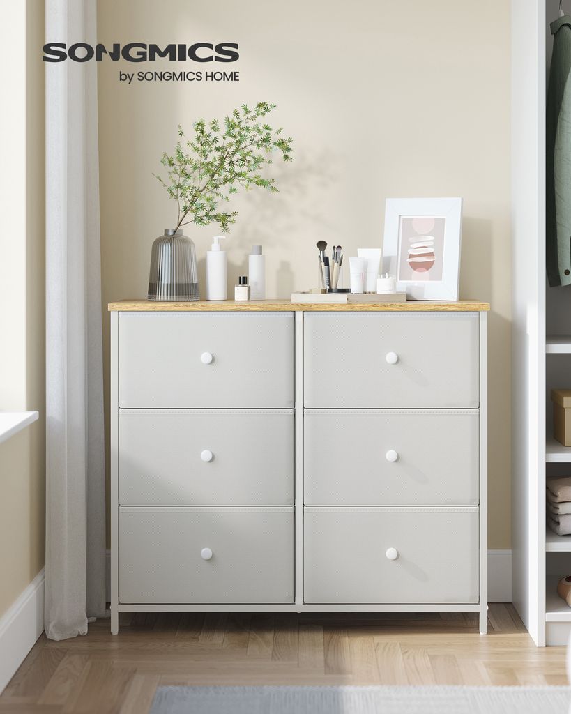 A dresser with drawers against a wall, decorated with a vase of plants, makeup brushes, and a framed picture.