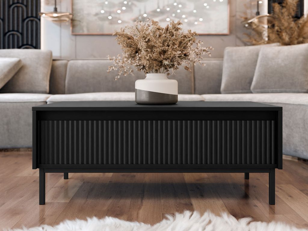 A black coffee table with a ribbed front is placed in a living room setting, in front of a sofa. A vase with dried flowers sits on top of the table.