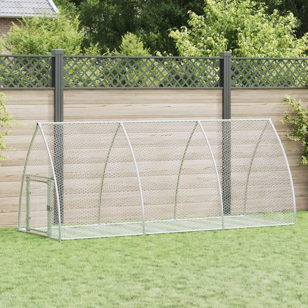 A metal chicken coop is set up on a grassy lawn in front of a wooden fence and a decorative lattice fence.