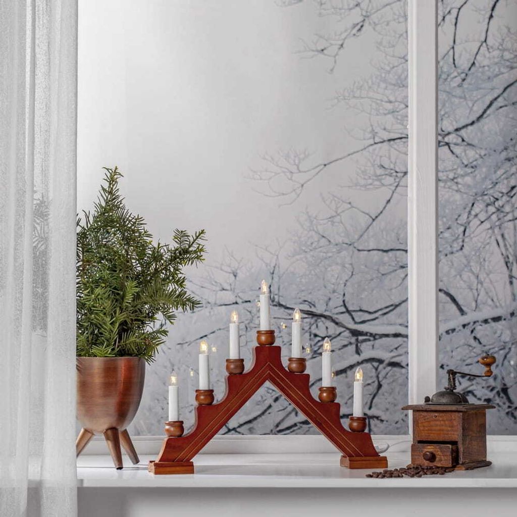 A wooden candle holder with seven electric candles is placed on a windowsill next to a small Christmas tree and a coffee grinder.