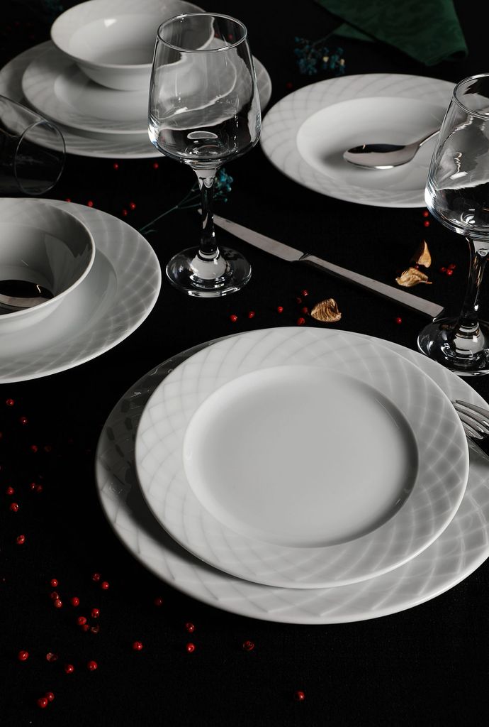 A formal table setting with white porcelain plates, bowls, and wine glasses on a black tablecloth, decorated with red peppercorns.