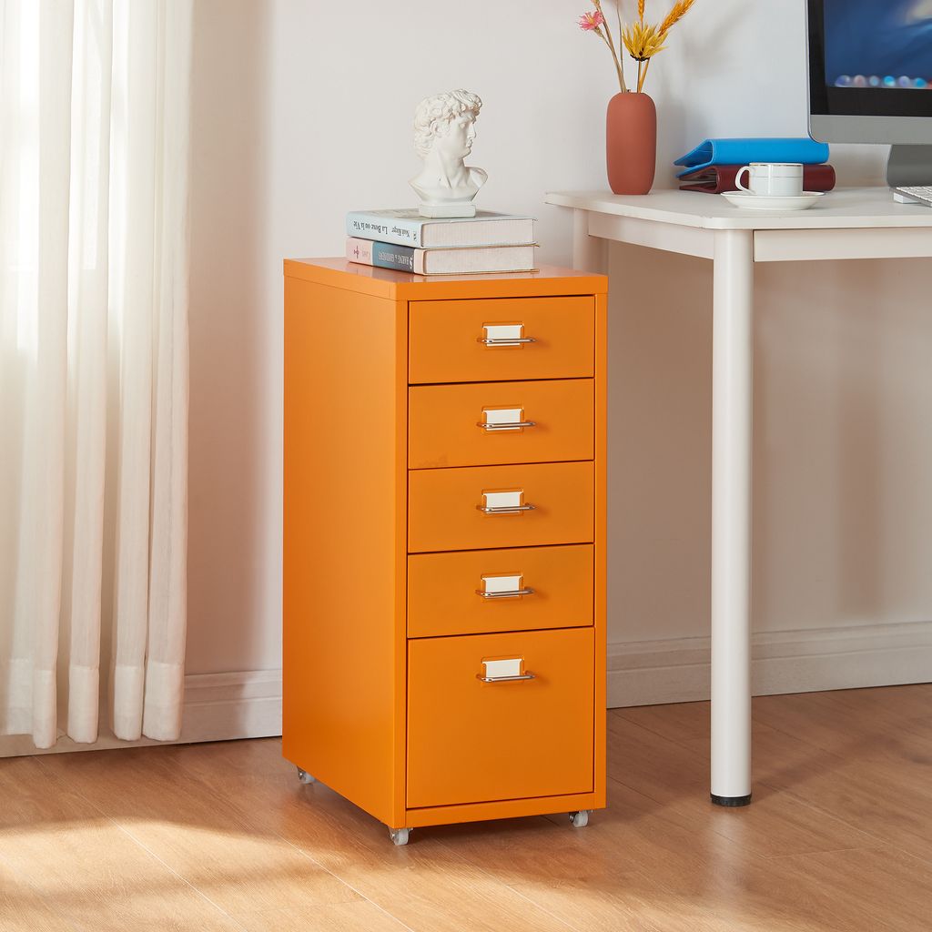 An orange filing cabinet with five drawers is placed next to a white desk. The cabinet has silver handles and is on wheels. There are books and a bust on top of the cabinet, and a vase with flowers and a computer monitor on the desk.