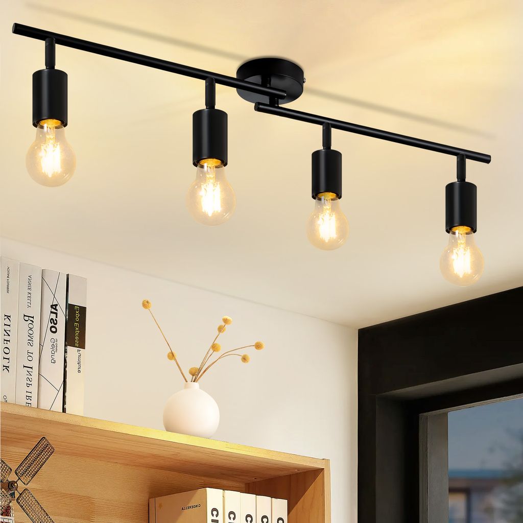 A black track light fixture with four exposed bulbs is mounted on a white ceiling above a wooden shelf with books and a small vase.