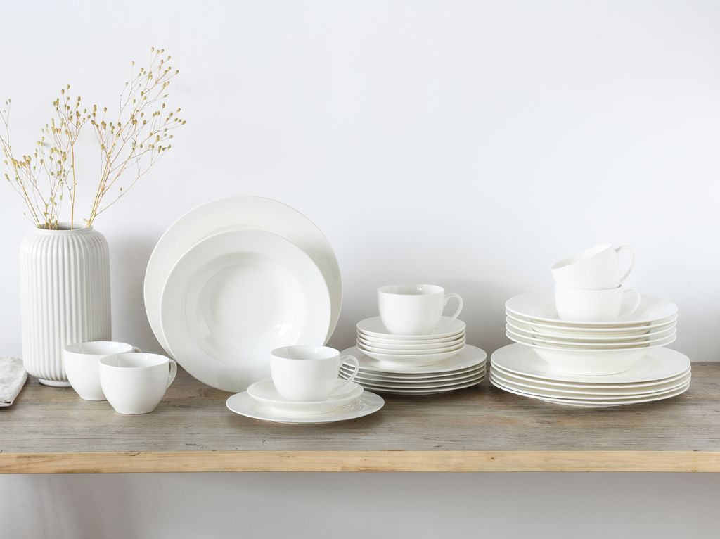 A collection of white porcelain dinnerware, including plates, bowls, and cups, arranged on a wooden shelf against a white background.