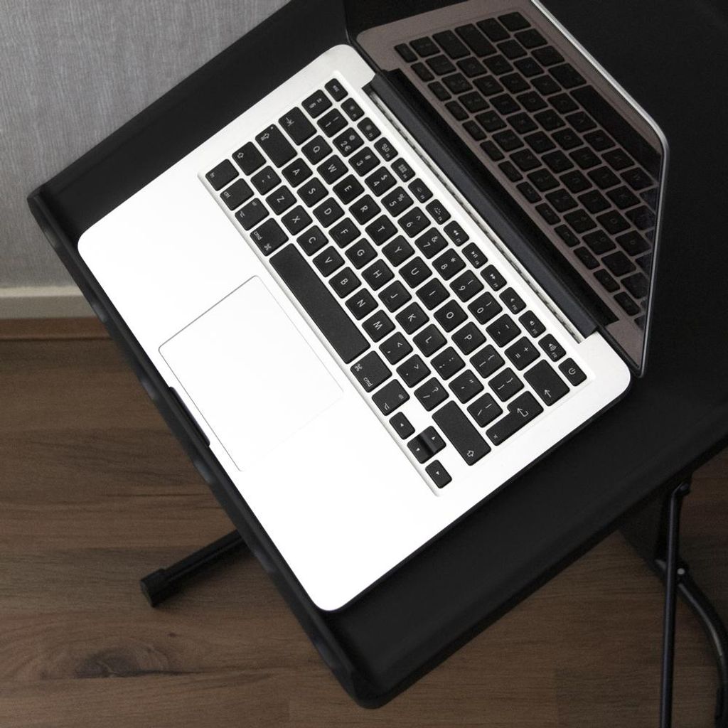 A laptop is placed on a black folding table, which is on a wooden floor. The background is a gray wall.
