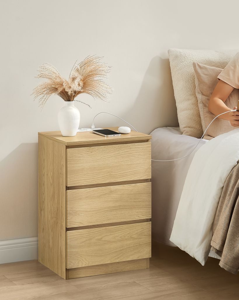A bedside table with a vase of dried flowers and a phone charging next to a bed.