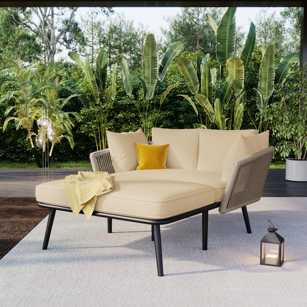 A beige outdoor sofa with an ottoman, a yellow throw pillow, and a yellow blanket is set up on a light-colored rug in a lush garden setting.