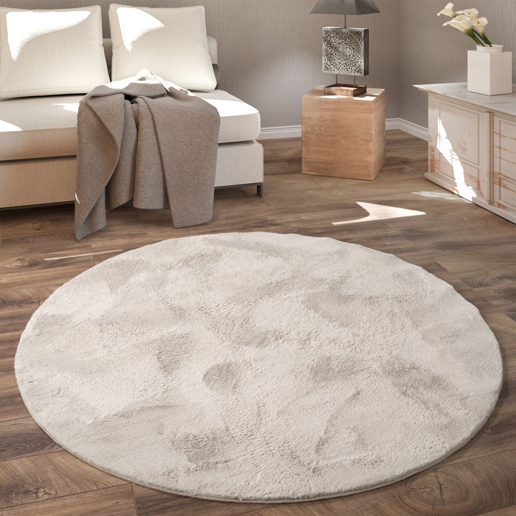 A round, fluffy rug is placed on a wooden floor in a living room setting, with a sofa and side table visible.