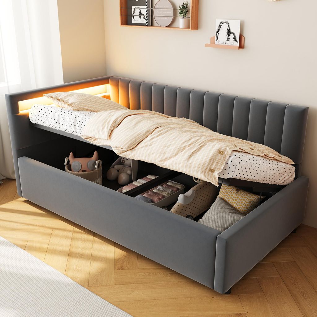 A grey upholstered bed with storage drawers open, showing stored items. The bed has a headboard with vertical tufting and integrated LED lighting.