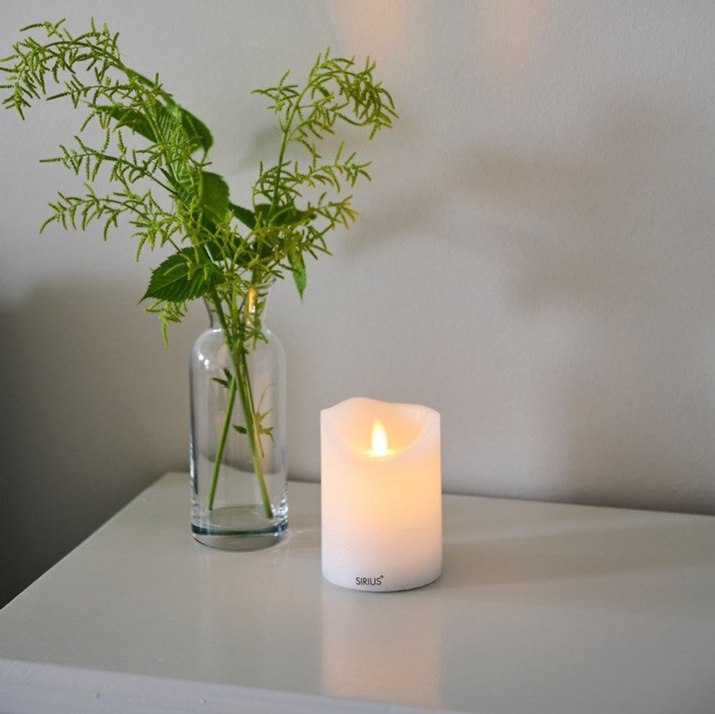 A white LED candle sits on a white surface next to a glass vase with green foliage.