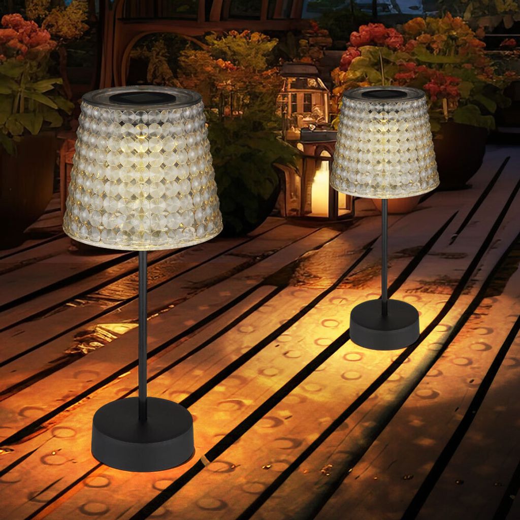 Two solar table lamps on a wooden deck at dusk, with plants and lanterns in the background.