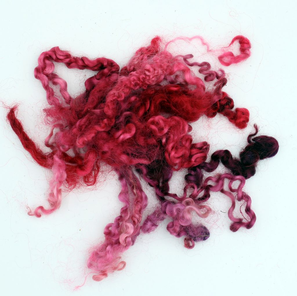 A pile of fluffy, curly wool fibers in various shades of pink, red, and purple, laid out on a white background.