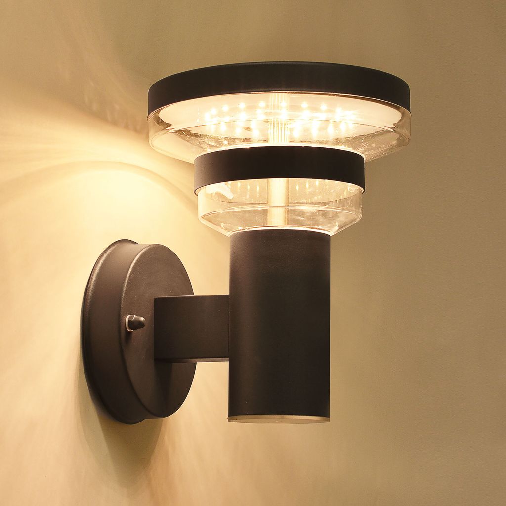 A modern black LED wall light with a clear glass shade is mounted on a light-colored wall.