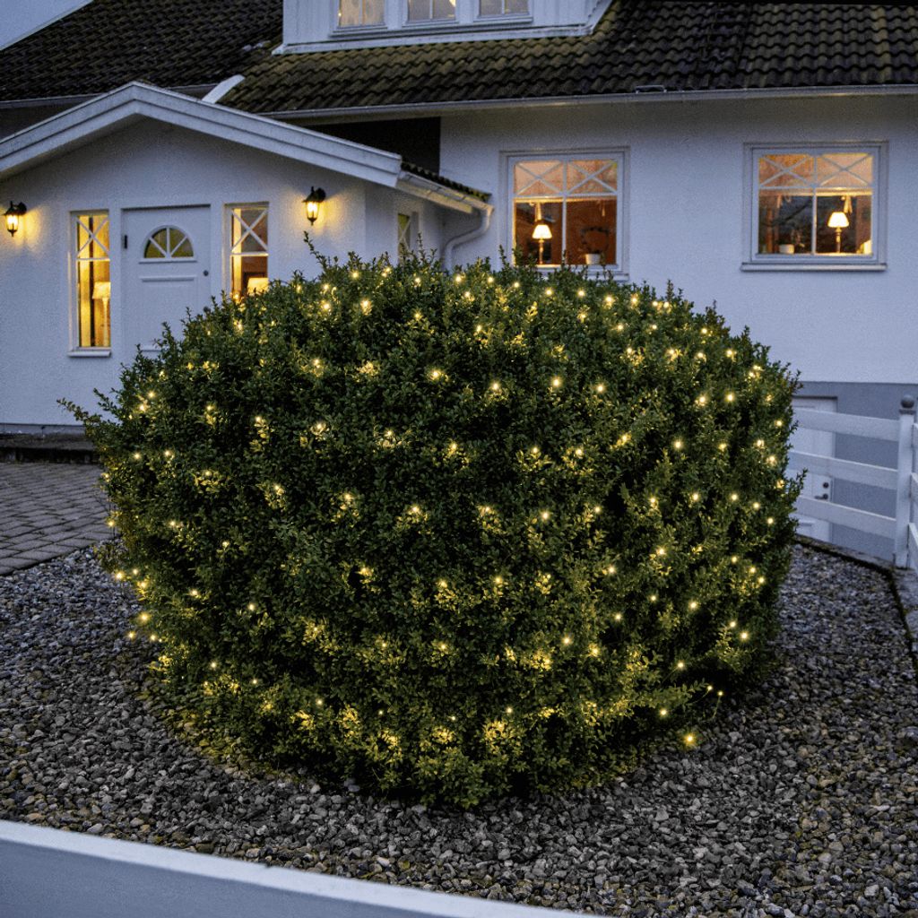 A bush decorated with LED lights in front of a house at dusk.