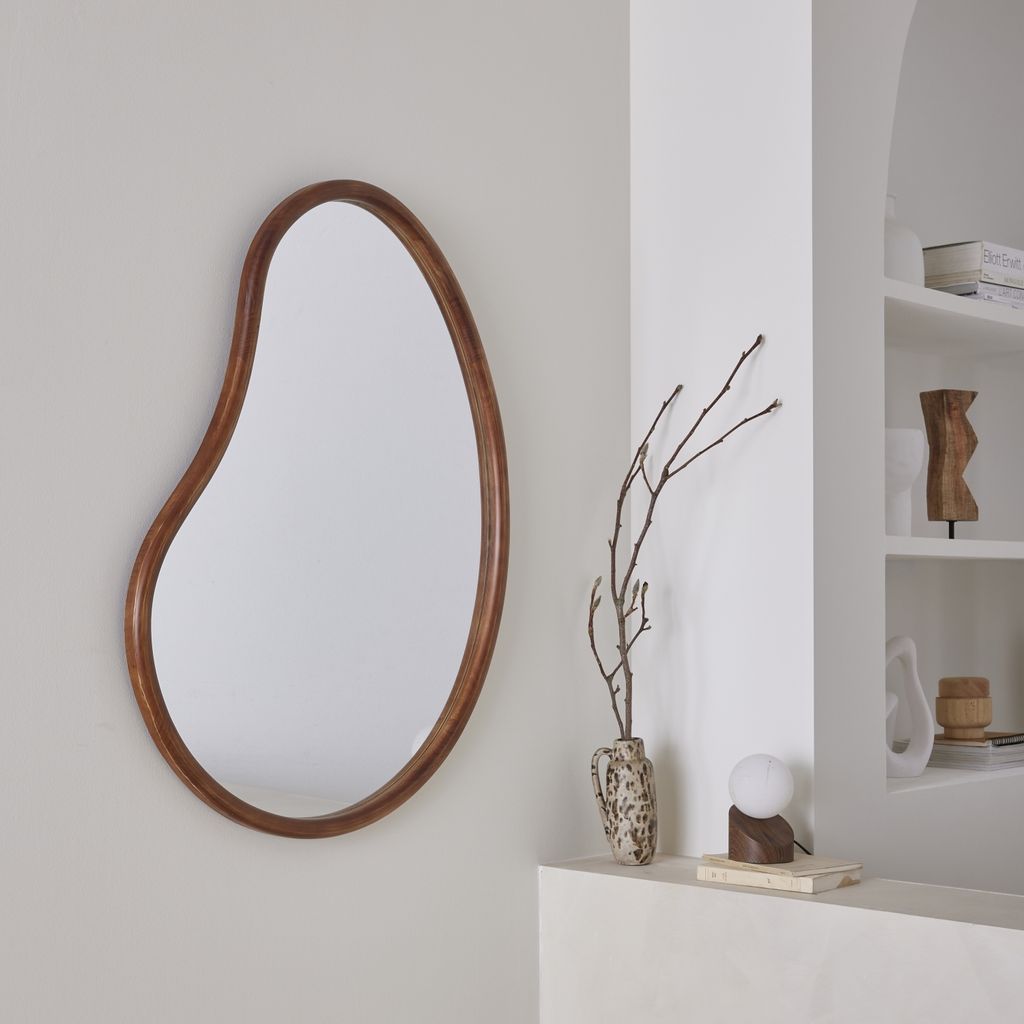 An organic-shaped mirror with a wooden frame hangs on a light gray wall next to a white shelf with decorative items.