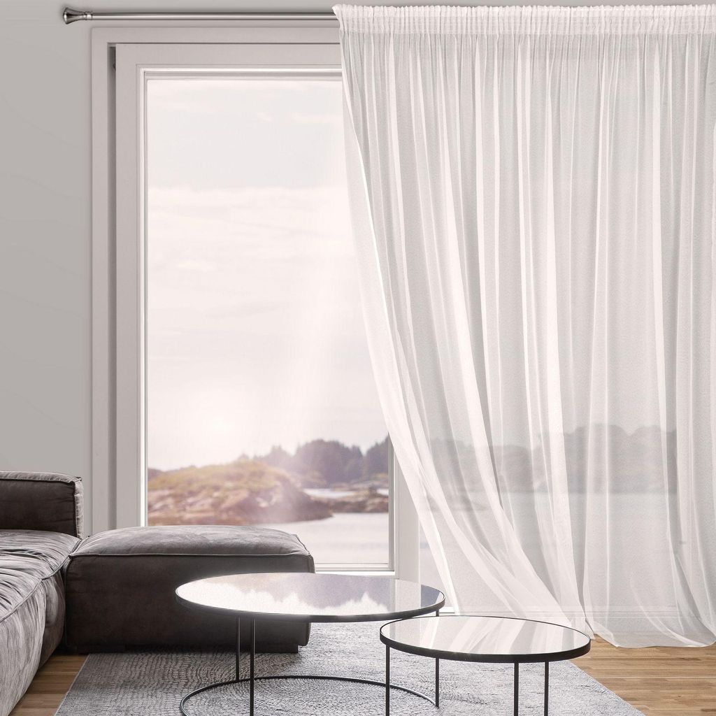 A sheer white curtain hangs in front of a large window overlooking a body of water and a rocky landscape. A sofa and ottoman are visible in the foreground.