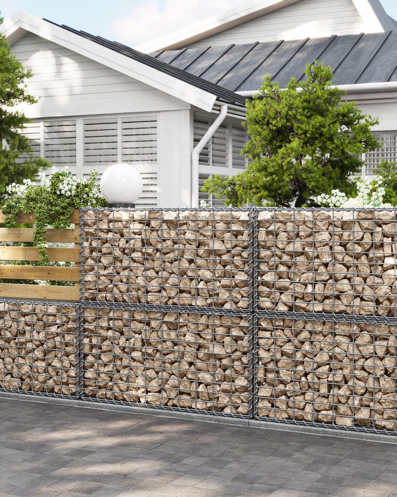 A gabion wall with a wooden fence section and a white sphere on top, in front of a white house with green trees.