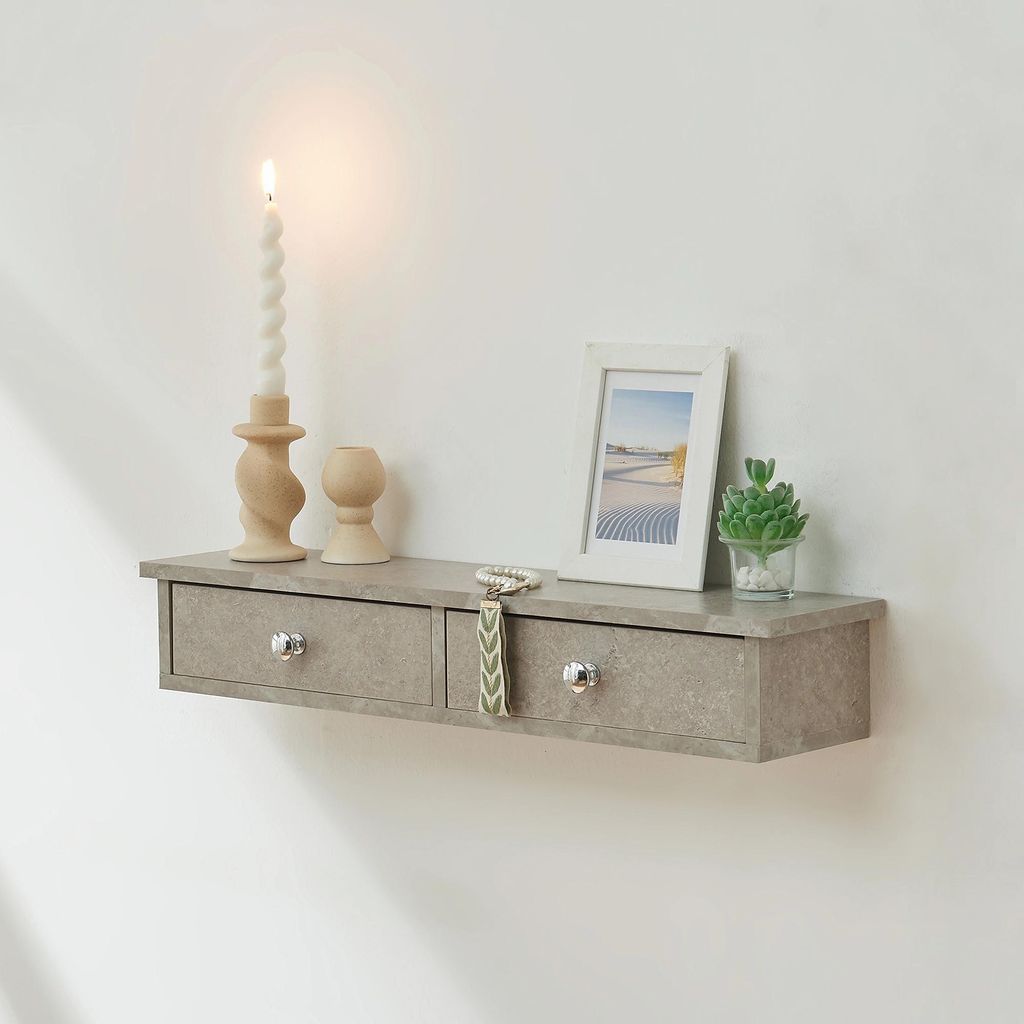 A floating shelf with two drawers mounted on a white wall, decorated with a candle, a picture frame, and a small plant.