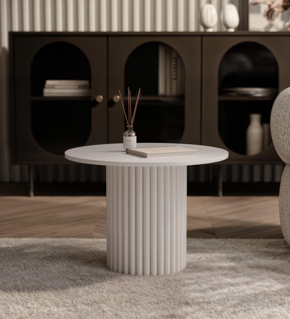 A round coffee table with a fluted base is placed on a rug in front of a dark cabinet.