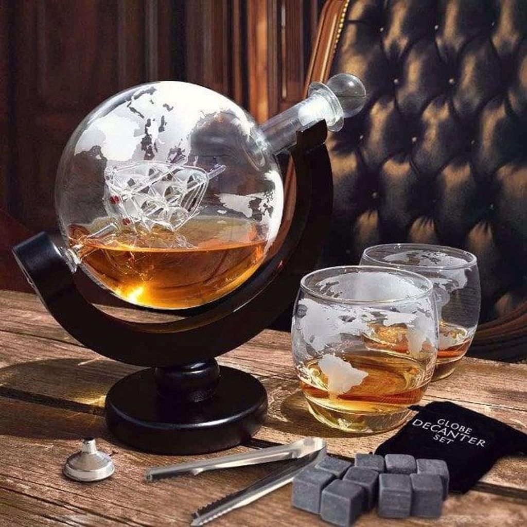 A globe-shaped decanter filled with amber liquid, two matching glasses, and whiskey stones on a wooden surface, with a tufted leather chair in the background.