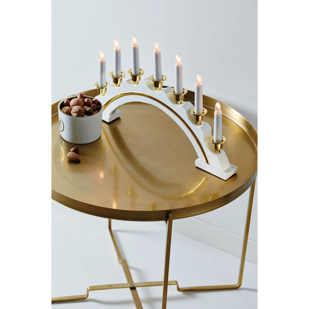 A white and gold candelabra with seven lit candles sits on a round, gold-colored table. A small white bowl filled with nuts is next to the candelabra, with a few nuts scattered on the table.